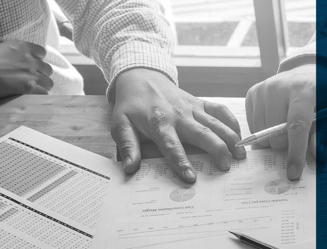 Black and white elegant professional photograph of hands of an accountant reviewing financial spreadsheets and documents with a luxury fountain pen, laptop and calculator nearby on a sleek wooden office desk. Cinematic depth of field, sharp focus, sophisticated corporate executive mood, clean aesthetic.